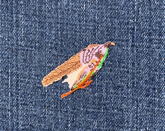 May include: An embroidered bird patch with brown, pink, and green details, and an orange and green accent. The patch is set against a blue denim background. The bird design is detailed.