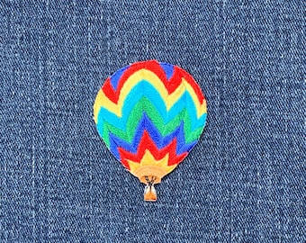 May include: A colourful hot air balloon patch with a vibrant design. The balloon features a red, yellow, blue, and green pattern. The patch is embroidered and has a small, gold-coloured basket attached.