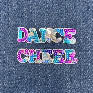 May include: Two iron-on patches featuring the words DANCE and CHEER in a bold, block font. The letters are covered in iridescent, multi-colored sequins that shift between shades of bright pink, vibrant blue, and silver, creating a reflective surface.