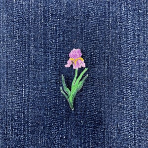 May include: An embroidered patch featuring a purple iris flower with a yellow center and green leaves, stitched onto a dark blue denim fabric background. The floral design displays detailed thread work in shades of violet and green.