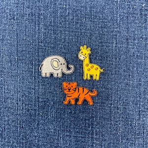 May include: Three embroidered fabric patches featuring cartoon animals: a light gray elephant, a yellow giraffe with brown spots, and an orange tiger with black stripes. The patches are arranged in a cluster against a dark blue denim fabric background.