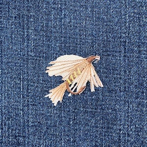 May include: Embroidered patch of a fly fishing lure in shades of tan, brown, and yellow. The patch is on a blue denim background. The fly lure has detailed stitching, with a wing-like design and a curved hook.