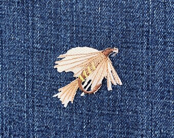May include: Embroidered fly fishing lure patch in tan, brown, and yellow hues. The patch is set against a blue denim backdrop. The fly lure features intricate stitching, with a wing-like shape and a curved hook.