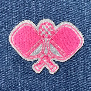 May include: A pink and silver embroidered patch in the shape of a pickleball paddle and ball. The patch has a glittery silver outline and is set against a blue denim background.