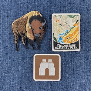 May include: Three embroidered patches: a brown and black bison, a rectangular Yellowstone National Park patch featuring a landscape with a blue river and yellow cliffs, and a square brown patch with a white icon of a pair of binoculars.