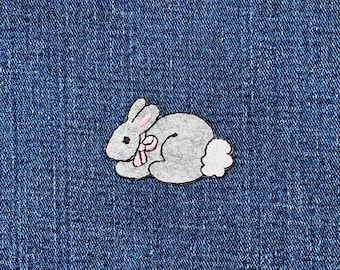 May include: A grey bunny patch with pink details, including the ears and a bow, and a white fluffy tail. The patch is set against a blue denim background.