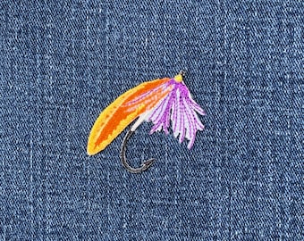 May include: Embroidered fishing fly patch with orange, yellow, and purple details, set against a blue denim background. The fly features a curved hook and is designed for anglers. This patch is suitable for jackets, bags, or other apparel.