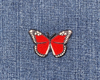 May include: A red butterfly patch with black and white detailing, displayed on a blue denim fabric. The embroidered design features a detailed butterfly, ideal for customising clothing or bags.