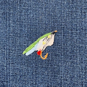 May include: Embroidered patch featuring a parrot with green, white, blue, red, and yellow details. The patch is set against a blue denim background, highlighting the bird's intricate design and vibrant colours.