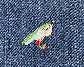 May include: Embroidered patch featuring a parrot with green, white, blue, red, and yellow details. The patch is set against a blue denim background, highlighting the bird's intricate design and vibrant colours.