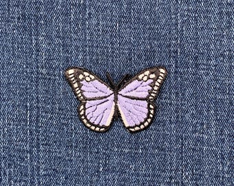 May include: A lavender butterfly patch with black and white embroidered detailing. The butterfly is set against a blue denim background. The patch has detailed wing patterns and a delicate design, suitable for clothing or accessories.