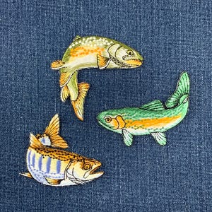 May include: Three embroidered fish patches featuring detailed stitching. The top fish is yellow and green with orange spots. The middle fish is green with an orange stripe. The bottom fish is white and yellow with blue vertical stripes.