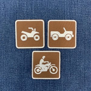 May include: Three square embroidered patches with brown backgrounds and white borders. The patches feature white stitched icons of an all-terrain vehicle, a four-wheeled off-road vehicle, and a person riding a motorcycle, all arranged on denim.