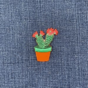 May include: Embroidered patch of a green cactus with red flowers in an orange pot with a green rim. The patch is on a blue denim background. This decorative patch is ideal for adding to clothing or accessories.
