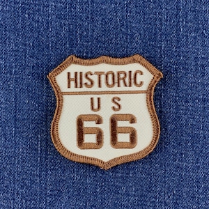 May include: A shield-shaped embroidered patch featuring a cream-colored background with a brown border. The text reads HISTORIC US 66 in brown lettering, styled like a road sign. The patch is attached to a textured blue denim fabric background.