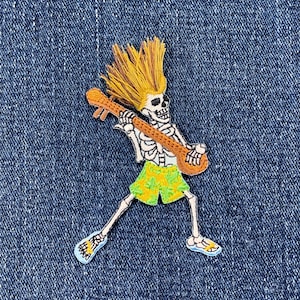 May include: Embroidered patch of a skeleton playing a ukulele. The skeleton has yellow hair and wears green and yellow shorts. The ukulele is brown. The patch is set against a blue denim background.