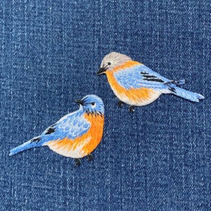 以下が含まれることがあります： 青い鳥の刺繍パッチ2つ。青、オレンジ、グレーのディテールが施されています。鳥は、質感のある濃い青色のデニムの背景に配置されています。パッチは、鳥の羽と形を示す詳細なステッチが特徴です。