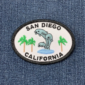 May include: White embroidered patch with black trim, featuring the text "SAN DIEGO CALIFORNIA" and a graphic of three dolphins jumping out of the water with palm trees on either side.