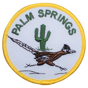 May include: A white embroidered patch with a yellow border. The patch features a roadrunner running across a desert landscape with a cactus in the background. The text "PALM SPRINGS" is embroidered in green around the border.