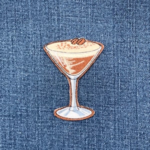 May include: Embroidered patch depicting a cocktail in a martini glass. The drink has a creamy, light-coloured top and a brown base, garnished with two coffee beans. The patch has a white outline and is set against a blue denim background.