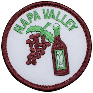May include: A white embroidered patch with a maroon border. The patch features a bottle of wine with a green label that says "Wine" and a bunch of grapes with green leaves. The text "Napa Valley" is written in a green cursive font around the bottle and grapes.