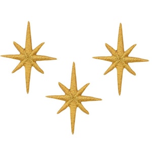 May include: Three gold star-shaped iron-on patches. Each star has eight points and is a bright gold color.