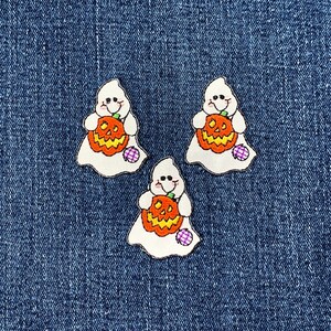 May include: Three Halloween-themed patches featuring white ghosts holding orange jack-o'-lanterns. Each ghost has a smiling face and is outlined in black. The pumpkins have yellow and orange details. A purple and white patch is at the bottom of each ghost.