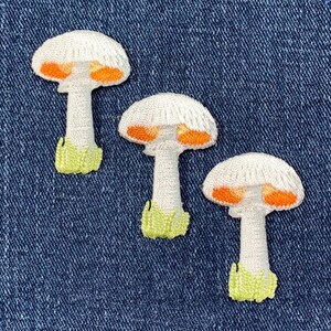 May include: Three white mushroom-shaped embroidered patches with orange accents. The patches are on a blue denim background.