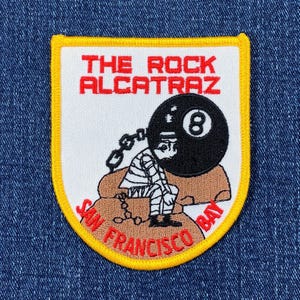 May include: A yellow and red embroidered patch with the text "The Rock Alcatraz San Francisco Bay" and a cartoon image of a prisoner with a ball and chain.