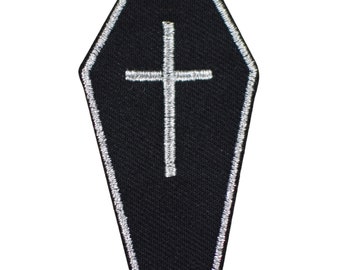 May include: A coffin-shaped embroidered patch featuring a black fabric base with a white stitched border. Centered on the patch is a white embroidered cross. The design is flat and symmetrical, showcasing clean lines and detailed thread work throughout.