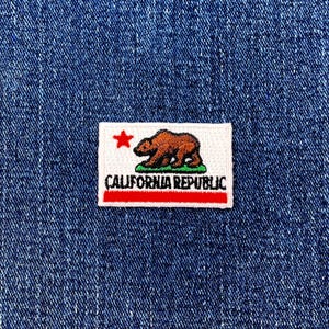May include: Embroidered patch of the California Republic flag, with a brown bear, red star, and text on a white background. The patch has a red border and green grass. The patch is on a blue denim background.