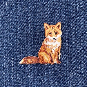 May include: Embroidered patch of a fox in shades of orange and white, sitting on a blue denim background. The fox has a fluffy tail. The patch is designed for application on clothing or accessories.