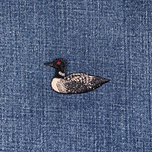 May include: An embroidered loon bird patch, with black, white, and grey plumage, a black head, and red eyes. The patch is set against a textured blue denim background, creating a contrast in textures and colours.