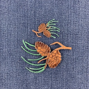 May include: Two embroidered patches featuring brown pinecones attached to light brown branches with thin, bright green pine needles. The patches are displayed against a dark blue denim fabric background with a visible woven texture.