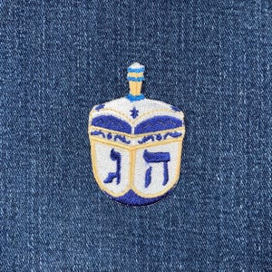 May include: A blue and white embroidered patch of a dreidel, a traditional spinning top played during Hanukkah. The dreidel has the Hebrew letters נ ג ש ה on its sides.