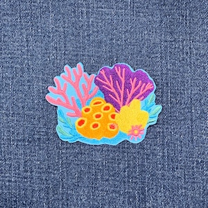 May include: An embroidered patch featuring a vibrant underwater coral reef scene. The design includes pink branching coral, a purple fan coral, a textured orange sponge with red spots, and a yellow sponge, all set against a light blue background.