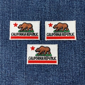May include: Three embroidered patches featuring the California Republic flag design. Each patch has a white background with a brown bear, a red star, and the words "CALIFORNIA REPUBLIC" in black. A red stripe runs along the bottom.
