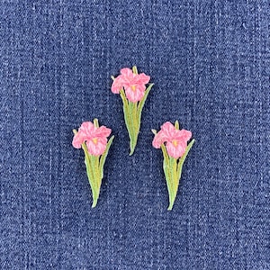 May include: Three embroidered patches featuring pink iris flowers with yellow centers and green stems and leaves. Each patch displays detailed stitching of the petals and foliage, arranged in a triangular formation against a dark blue denim fabric.