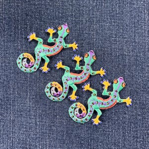 May include: Three green, blue and orange embroidered gecko lizards with rhinestones on a blue denim background.
