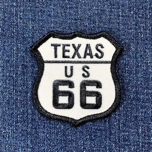 May include: A white and black embroidered patch in the shape of a Texas highway sign. The patch features the words "TEXAS", "US", and the number "66". The patch is set against a blue denim background.