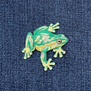 May include: Embroidered frog patch in shades of green, yellow, and cream, with detailed stitching. The frog is depicted in a relaxed pose, with its legs and webbed feet extended. The patch is set against a dark blue denim background.
