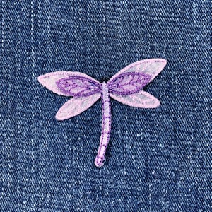 May include: A detailed dragonfly patch in lavender and pink, featuring delicate lace-like patterns on the wings. The patch is set against a textured, dark blue denim backdrop, offering a contrast in colour and texture.