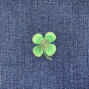 May include: A green four-leaf clover patch embroidered on a blue denim background. The clover has a detailed, textured appearance, with varying shades of green. The denim fabric provides a contrasting backdrop.