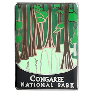 May include: A rectangular enamel pin featuring a forest scene with tall trees and a body of water. The pin has a black border and the words "CONGAREE NATIONAL PARK" in white text at the bottom.