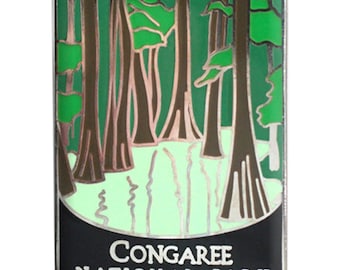 May include: A rectangular enamel pin featuring a forest scene with tall trees and a body of water. The pin has a black border and the words "CONGAREE NATIONAL PARK" in white text at the bottom.