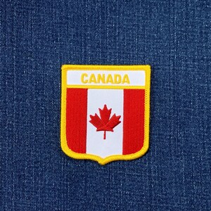 May include: A shield-shaped embroidered patch featuring the Canadian flag with a red maple leaf on a white vertical stripe flanked by two red stripes. The word CANADA is stitched in yellow on a white horizontal band at the top, bordered by yellow.