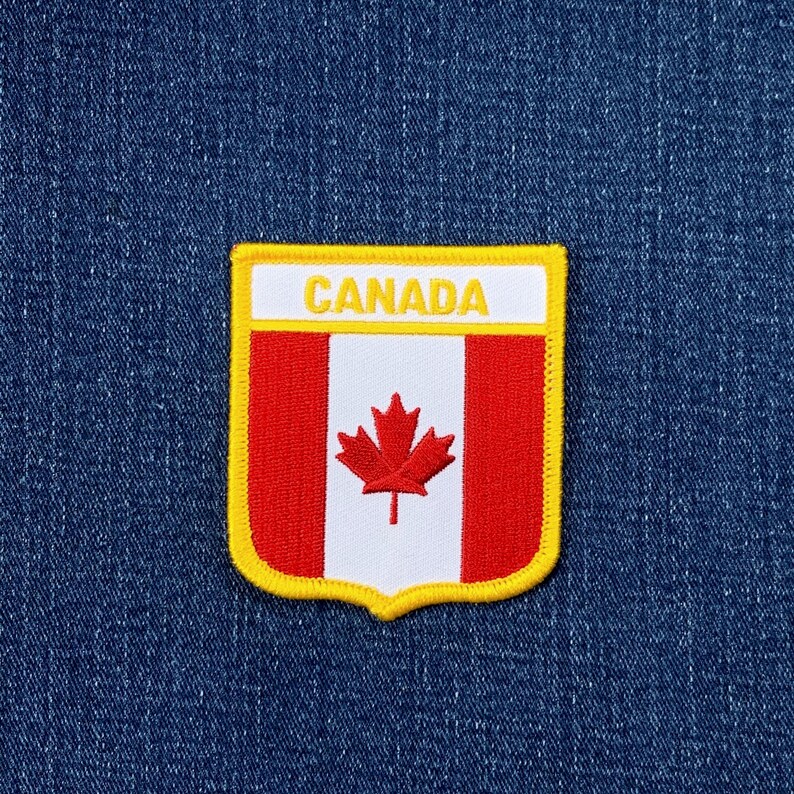 May include: A shield-shaped embroidered patch featuring the Canadian flag with a red maple leaf on a white vertical stripe flanked by two red stripes. The word CANADA is stitched in yellow on a white horizontal band at the top, bordered by yellow.