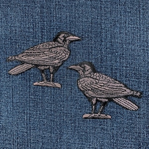 May include: Two embroidered raven patches. The ravens are in a standing position, with detailed feather patterns in shades of gray and black. The patches are set against a blue denim background.