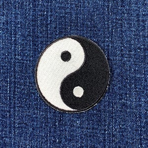 Peut inclure: Un écusson circulaire représentant le symbole Yin et Yang, en noir et blanc. L'écusson a une bordure noire et est posé sur un fond en denim bleu.