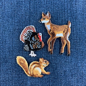 Peut inclure: Trois écussons en tissu brodé représentant un cerf brun debout, une dinde noire et blanche avec une caroncule rouge, et un écureuil brun à la queue touffue. Chaque écusson présente un travail de fil détaillé représentant les animaux.